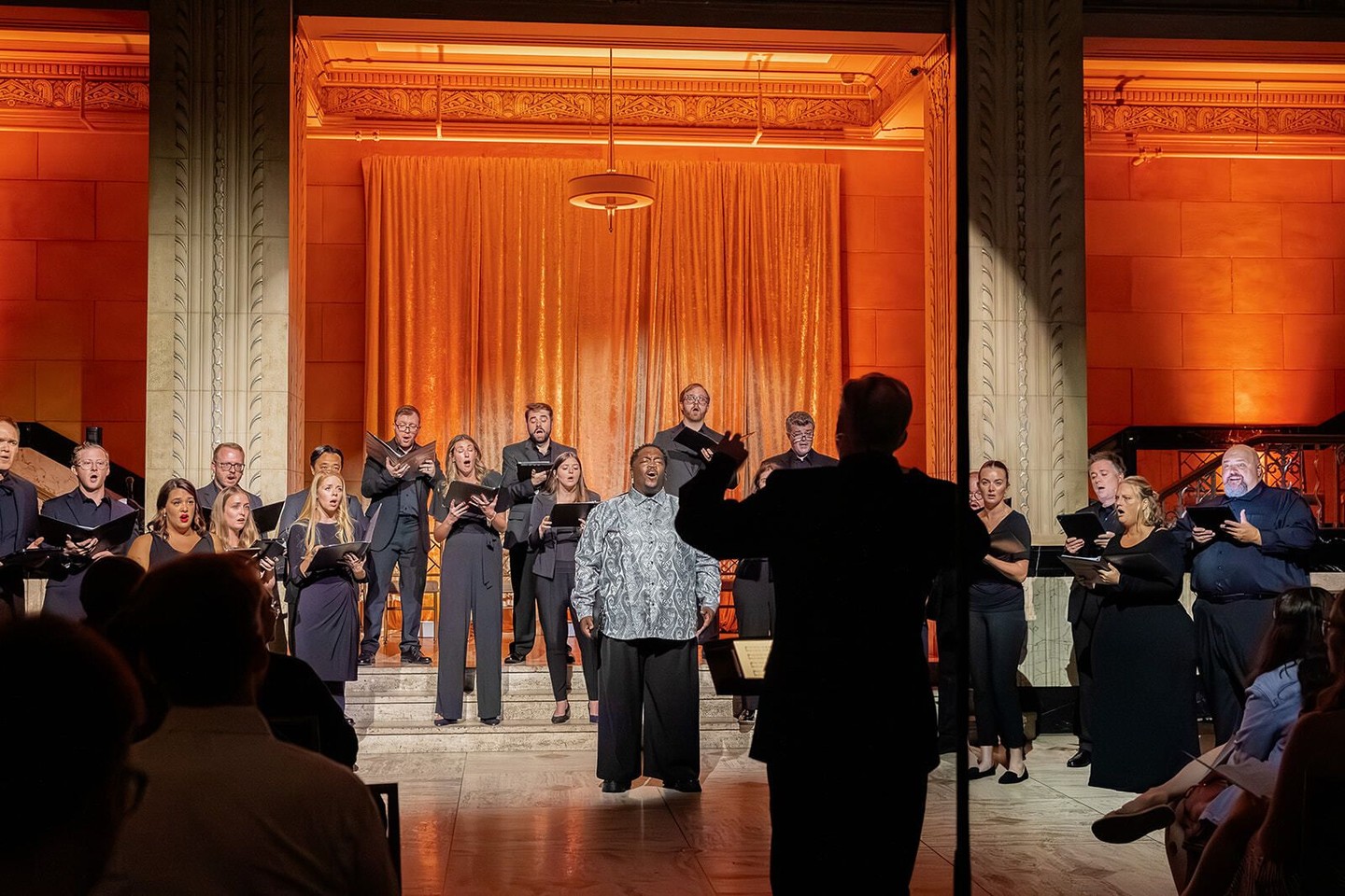 Joshua Oppenheim conducting a choir on stage in a warmly lit concert hall